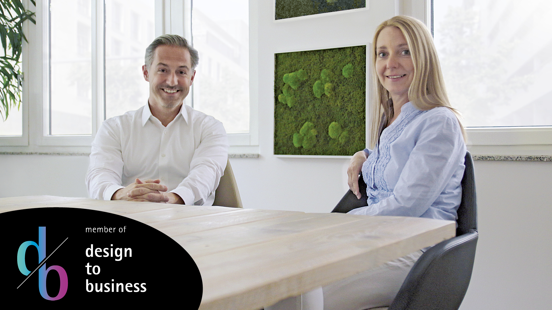 Foto von zwei Personen, eine Frau und ein Mann, sitzen im Büro auf Stühlen und lächeln in die Kamera. Sie sind Mitglied im Design top business.