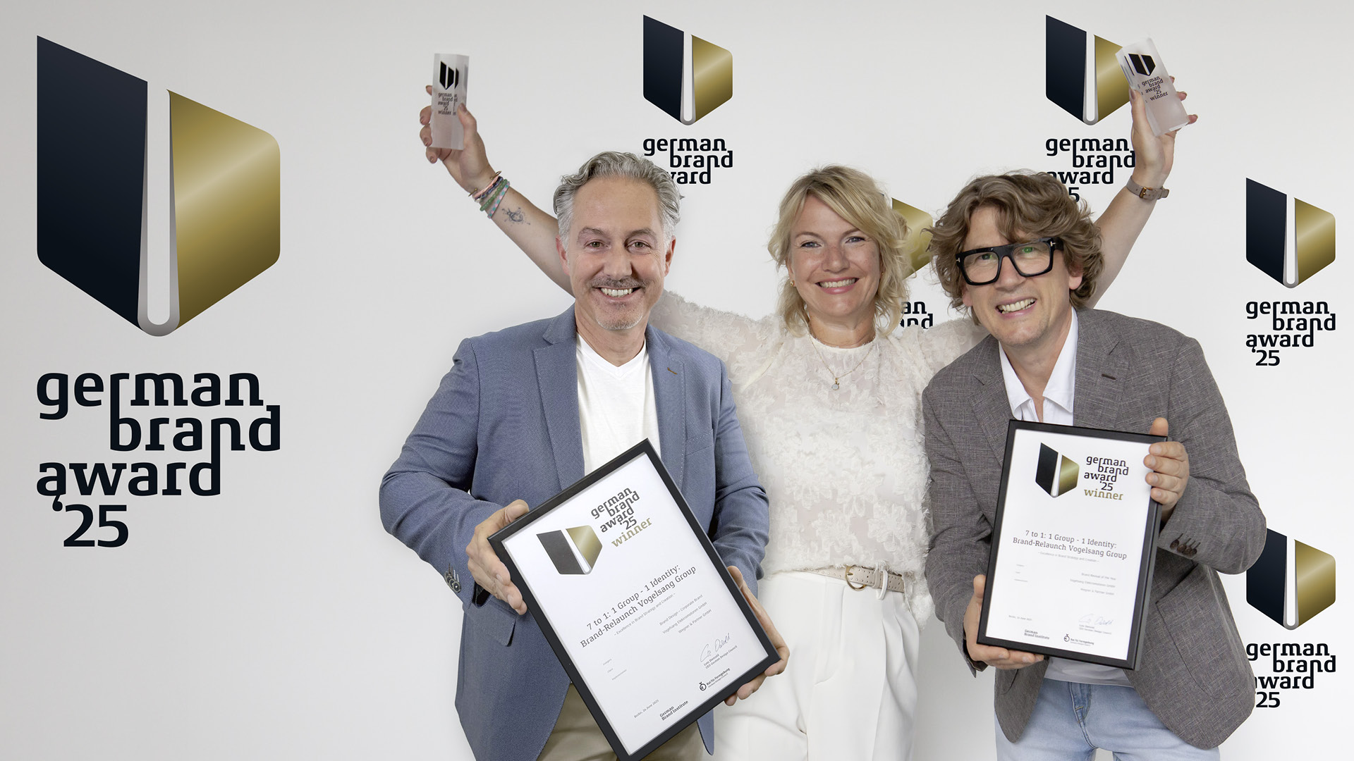 Drei Personen von wegner und partner stehen vor einer Fotowand mit dem Logo des German Brand Award 2025 und halten Urkunden und Trophäen in den Händen. Alle lächeln in die Kamera. Das Bild zeigt die Auszeichnung des Projekts „7 to 1: Group – 1 Identity / Brand Relaunch Vogelsang Group“.