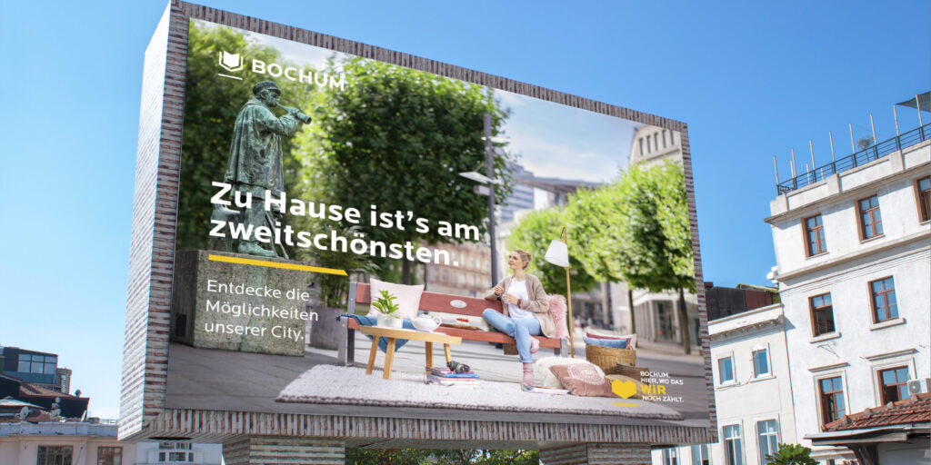 Großes Werbeplakat der Stadt Bochum an einer Häuserfassade mit dem Motiv „Zu Hause ist’s am zweitschönsten“, das eine Frau auf einer gemütlich eingerichteten Parkbank in der Innenstadt zeigt.