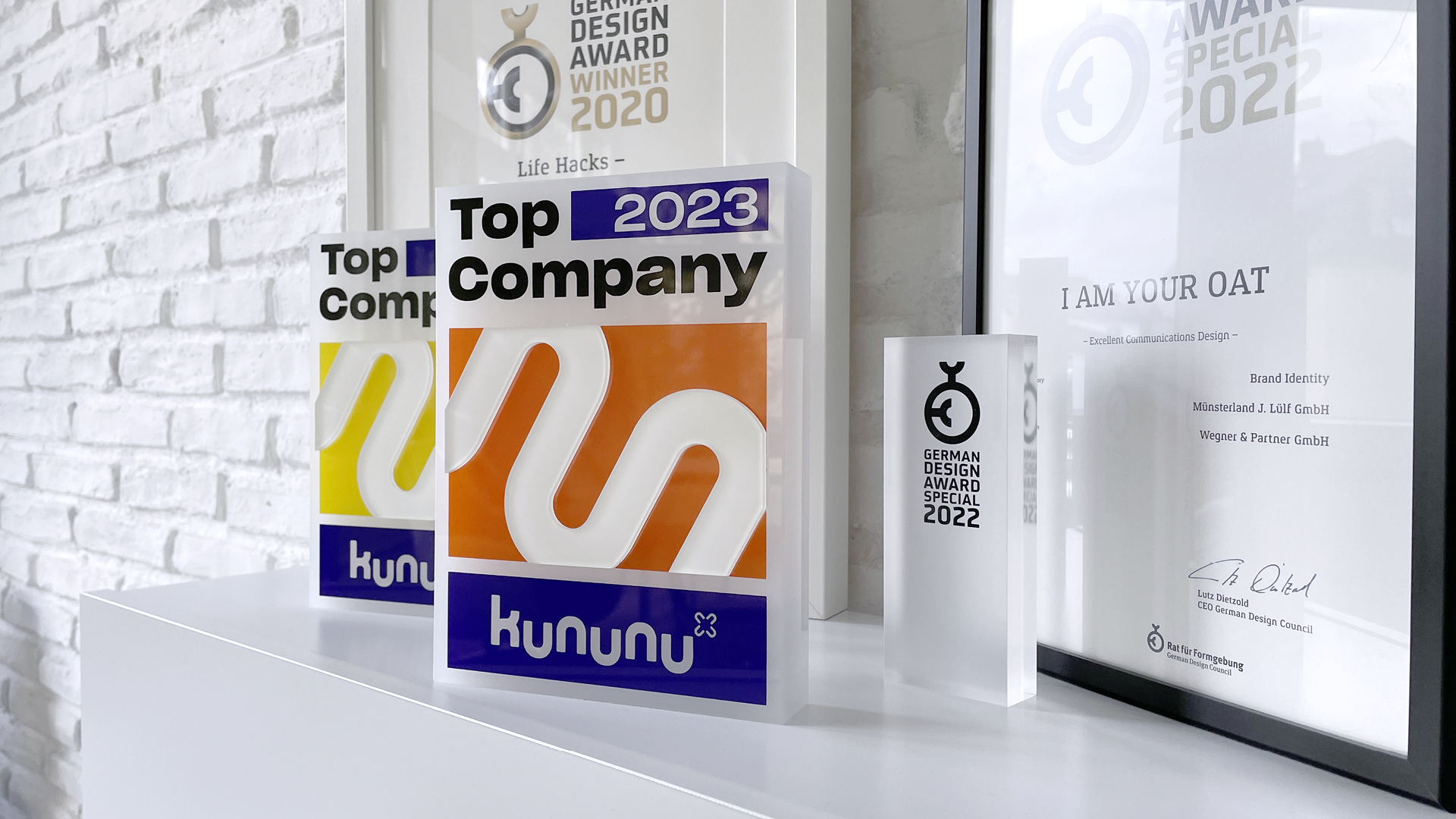 Mehrere Auszeichnungen auf einem weißen Sideboard vor einer hellen Backsteinwand. Im Vordergrund stehen Kununu-Trophäen „Top Company 2023“ und eine „German Design Award Special 2022“-Auszeichnung. Im Hintergrund sind gerahmte Urkunden zu sehen. Das Bild zeigt prämierte Awards von wegner & partner.
