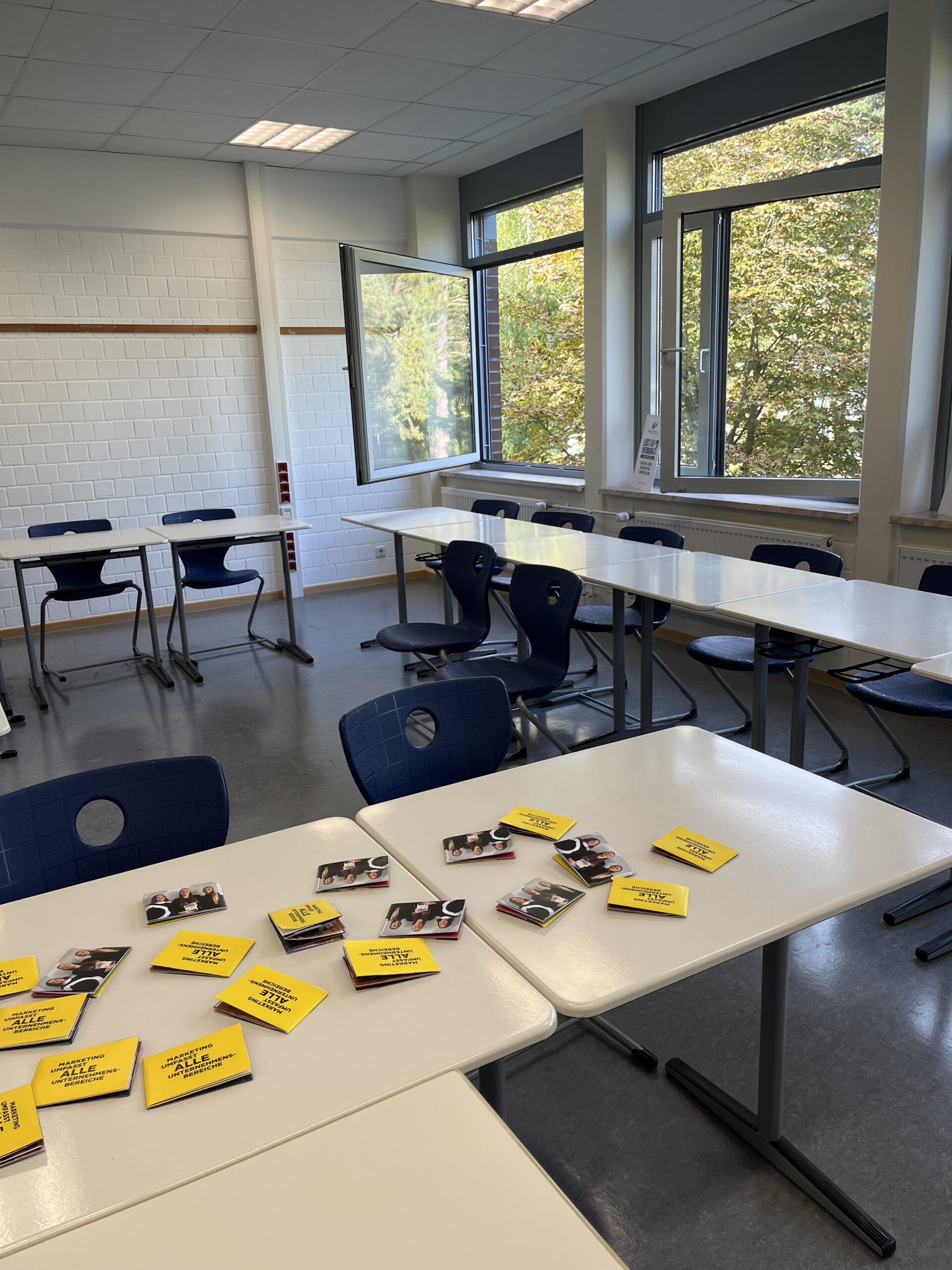 Blick in einen hellen Klassenraum mit weißen Tischen und blauen Stühlen. Auf den vorderen Tischen liegen mehrere Flyer von wegner & partner.