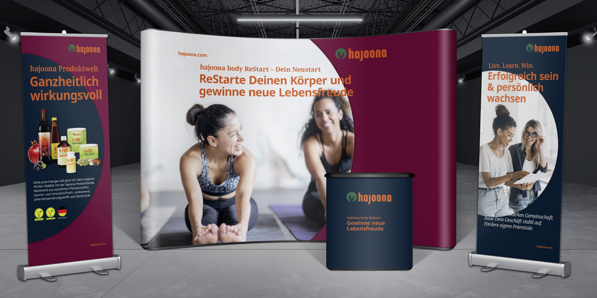 Messestand von hajooona mit gebogener Rückwand und zwei Roll-ups: links die Produktwelt mit Nahrungsergänzungsmitteln, in der Mitte das Programm „body ReStart“ mit sportlichem Motiv, rechts ein Banner zum Thema persönlicher Erfolg und Wachstum.