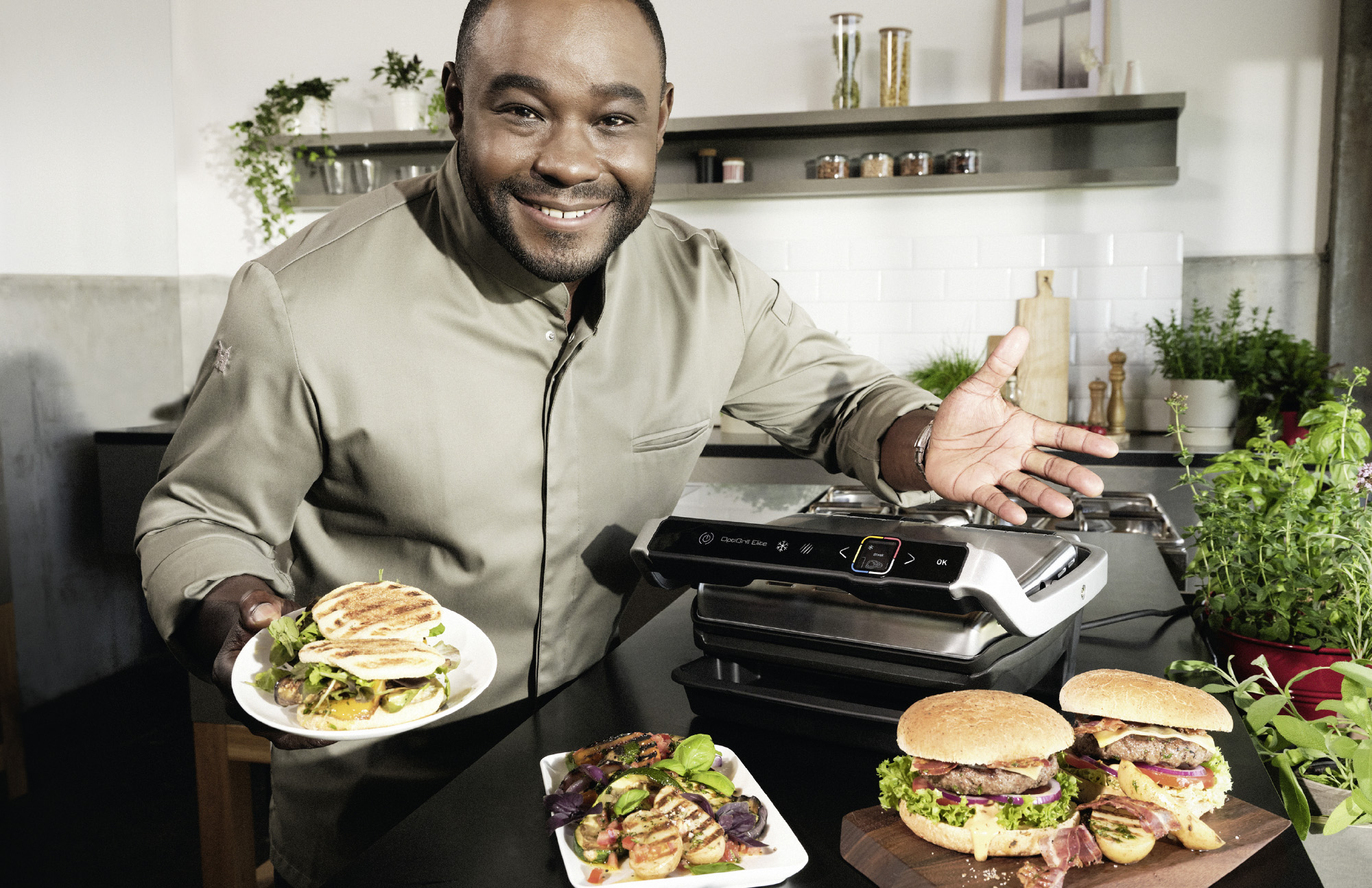 Nelson Müller präsentiert in einer modernen Küche verschiedene Gerichte – Steak, Burger, Pommes und Hähnchen – neben dem Opti-Grill von Tefal.