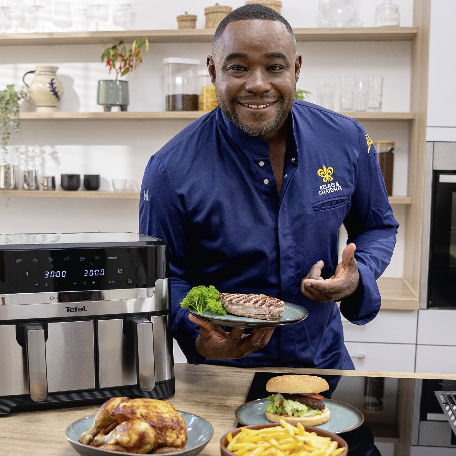 Nelson Müller präsentiert in einer modernen Küche verschiedene Gerichte – Steak, Burger, Pommes und Hähnchen – neben einer Heißluftfritteuse von Tefal.