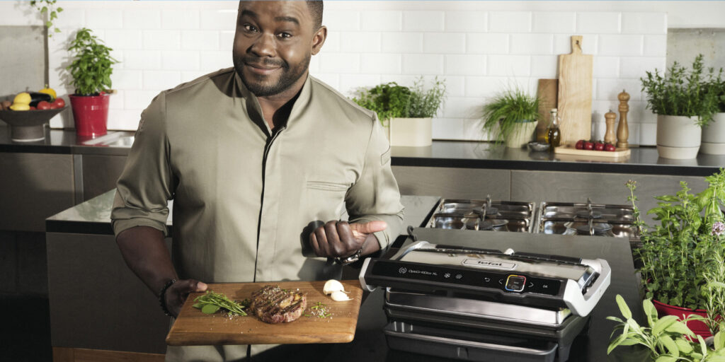 Nelson Müller steht in einer modernen Küche und präsentiert auf einem Holzbrett ein Steak mit Kräutern, neben ihm steht der Opti-Grill von Tefal.