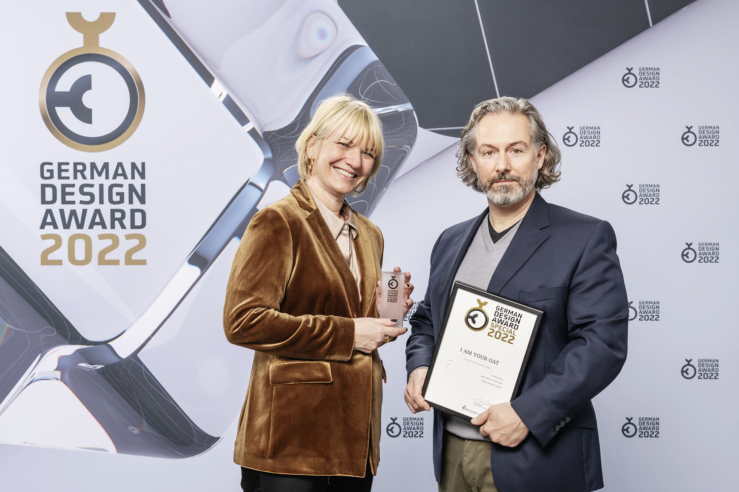 Zwei Personen, eine männliche und eine weibliche, stehen vor einer Fotowand des German design awards 2022 und halten eine Urkunde und ein Pokal in die Kamera. Die Frau auf dem Bild lächelt.