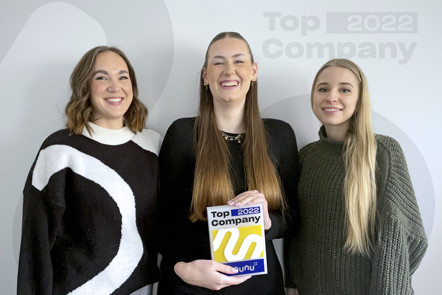 Drei Frauen stehen vor einer Wand neutralen Wand mit dem Logo Top Company 2022 von kununu. alle drei Personen lächeln in die Kamera.