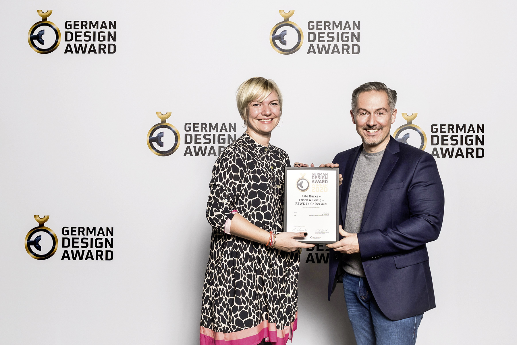 Zwei Personen stehen vor einer Fotowand mit dem Logo des German Design Award und halten gemeinsam eine Urkunde. Die Szene zeigt die Preisverleihung des German Design Award, bei der das Projekt „Life Hacks – Frisch & Fertig / REWE To Go bei Aral“ ausgezeichnet wurde. Beide Personen lächeln in die Kamera.